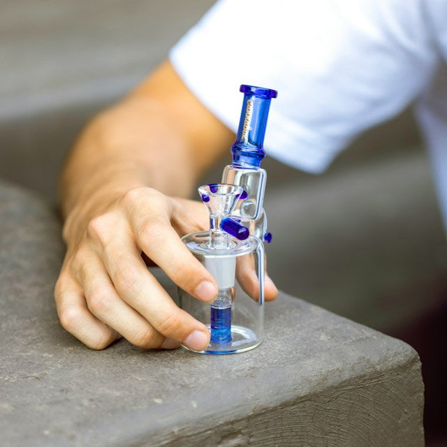 a man in a white t shirt with his hand holding a blue mini bong made from quality glass